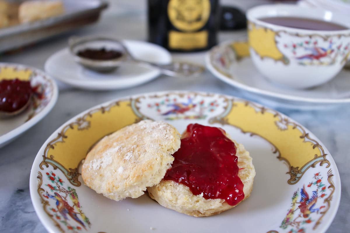 a tea scone topped with jam, served on china with a cup in the background