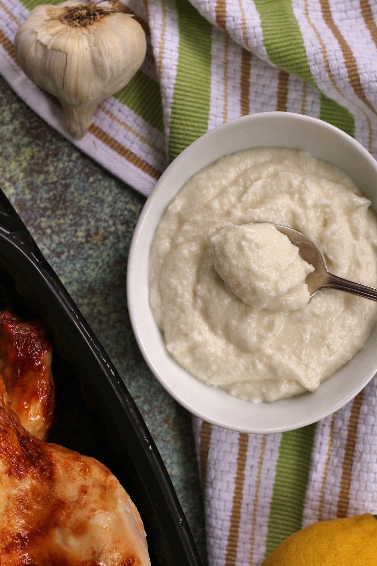 overhead view of garlic sauce in a white bowl next to chicken, lemon, and garlic