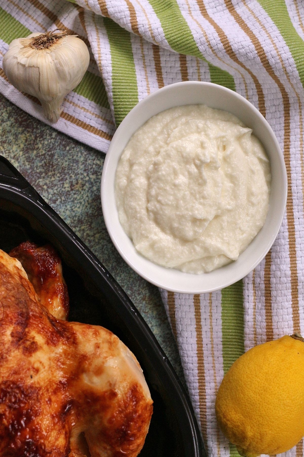 overhead view of a bowl of garlic sauce, head of garlic, rotisserie chicken, and lemon