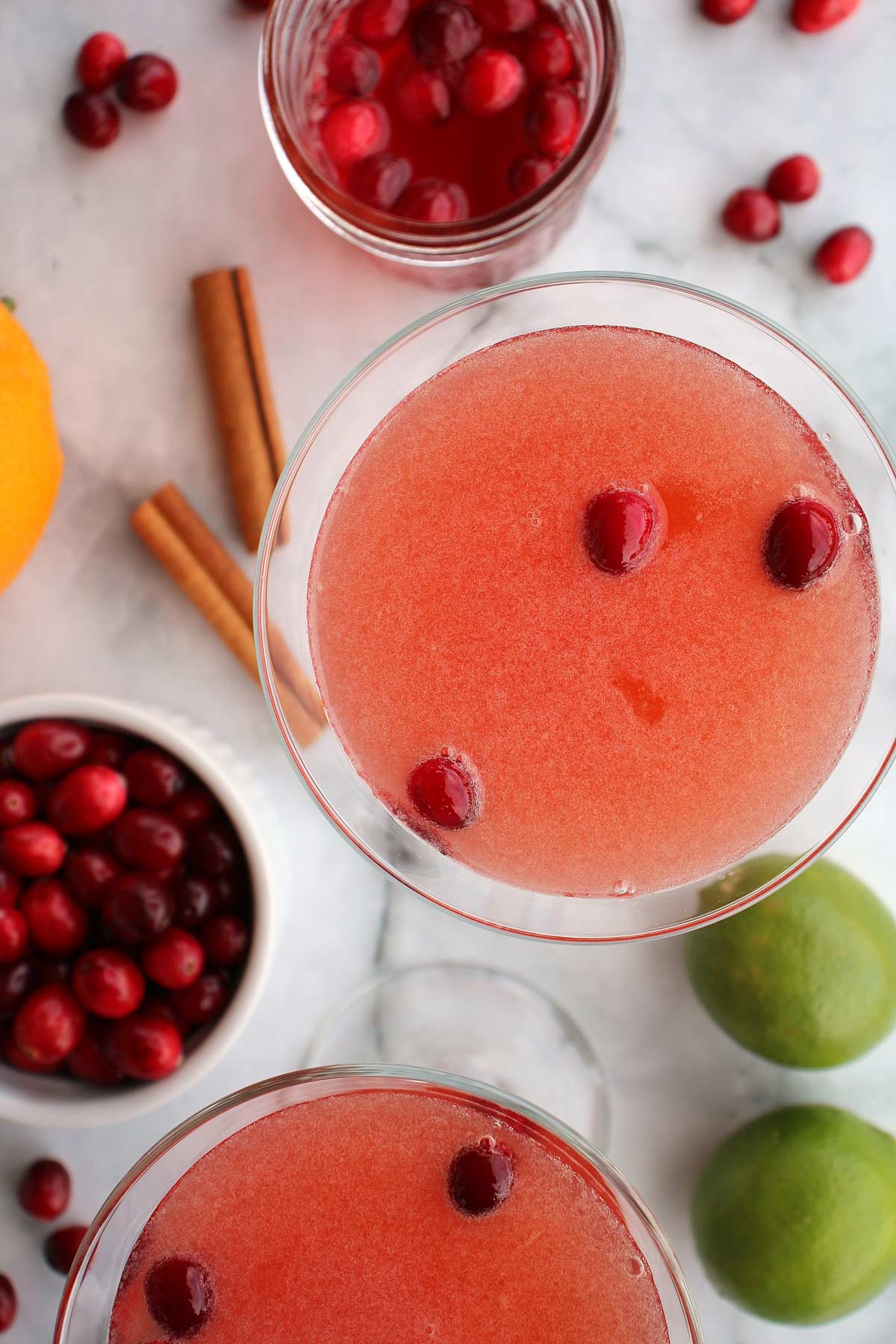 Two cranberry daiquiris, cranberries, cinnamon sticks, and limes on a marble surface.