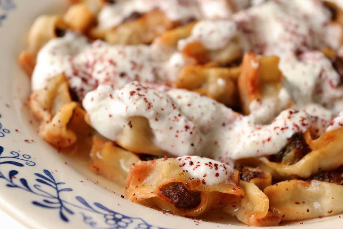 Closeup of a shallow white bowl filled with canoe-shaped dumplings topped with yogurt sauce.