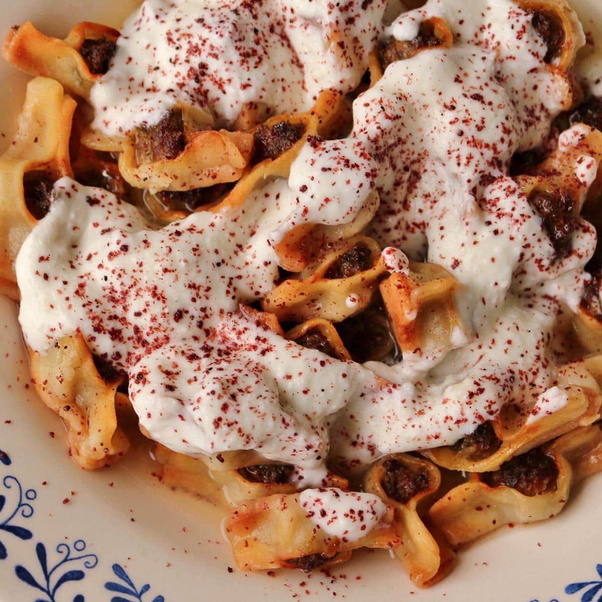 A shallow bowl filled with canoe-shaped meat-filled dumplings topped with yogurt sauce.