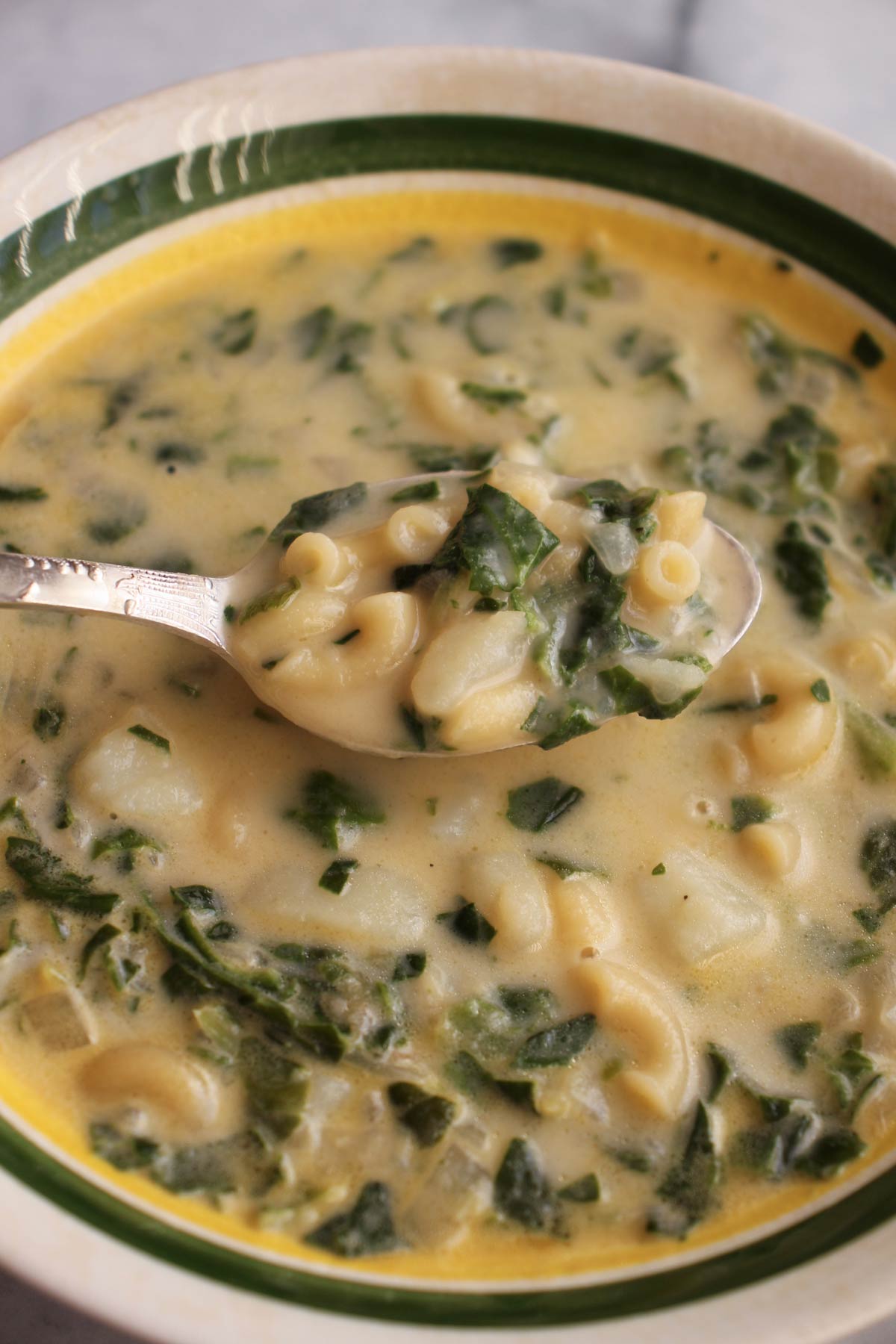 Closeup of a spoonful of soup with macaroni, potatoes and spinach lifting out of a bowl.