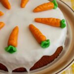 A glazed Swiss carrot cake topped with marzipan carrots on a rustic round serving dish.