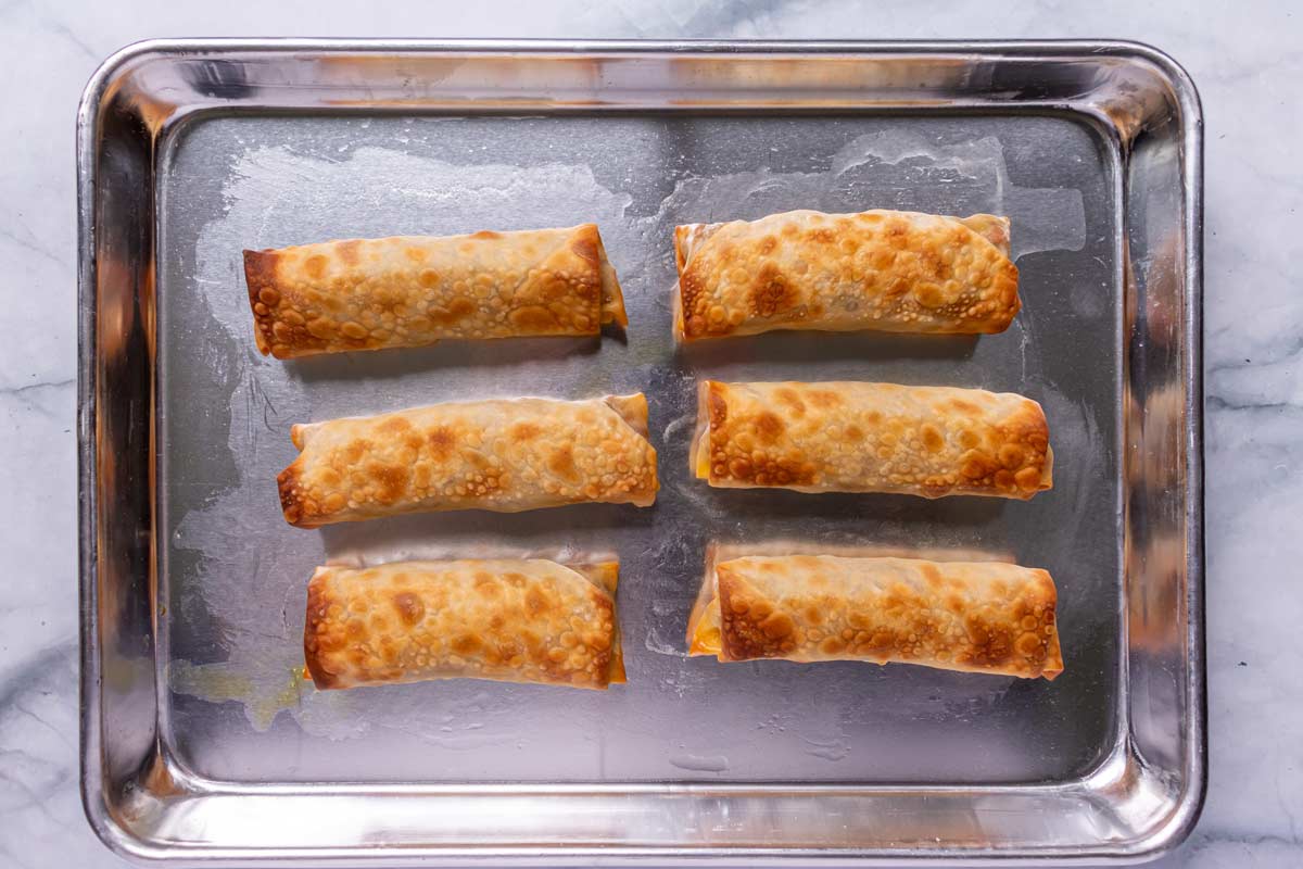 Six baked egg rolls on a small metal baking sheet.