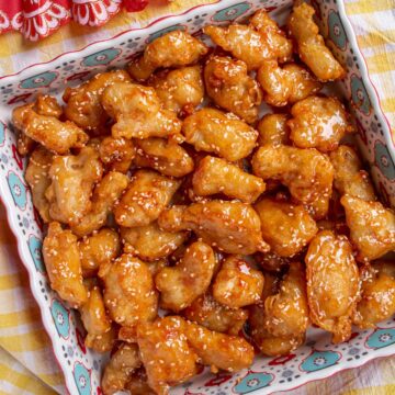 A colorful square dish filled with Chinese honey sesame chicken next to a red folding fan.
