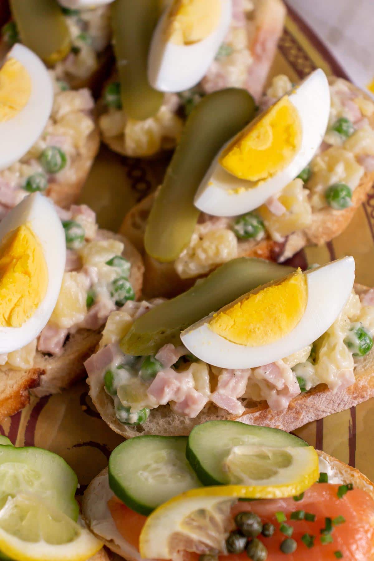 Closeup of open-faced sandwiches topped with potato and ham salad, sliced pickle and boiled egg.