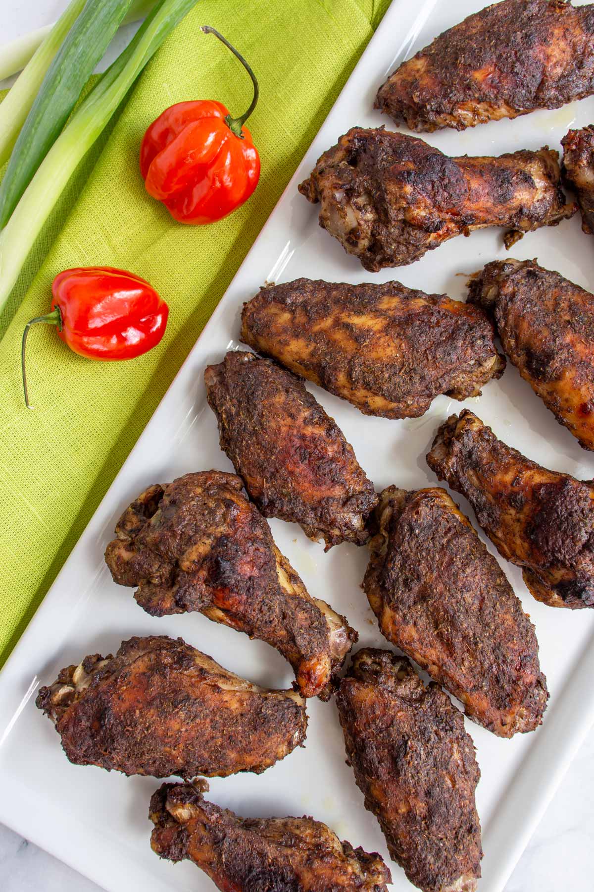 Closeup of Jamaican jerk chicken wings on a white rectangular plate next to 2 habanero peppers.