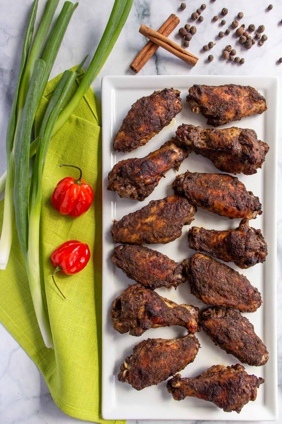 A white rectangular plate of jerk chicken wings next to scallions, chiles, cinnamon sticks and allspice berries.