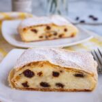 Two slices of cheese strudel with raisins topped with powdered sugar on small white plates.