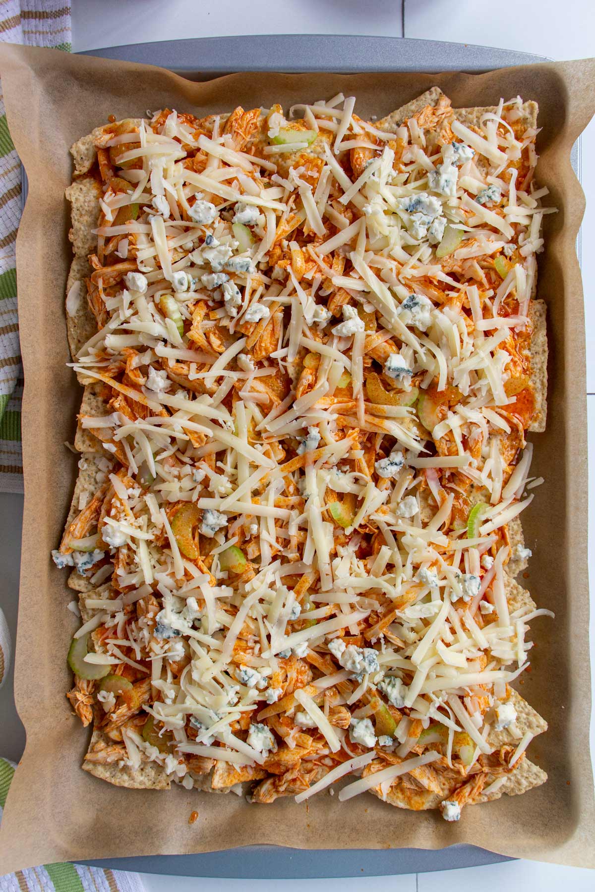 A sheet pan of Buffalo chicken nachos before baking with unmelted cheese on top.