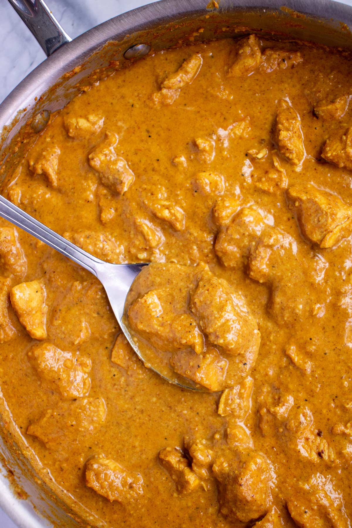 A stainless steel skillet of chicken korma curry with a large metal spoon scooping some up.