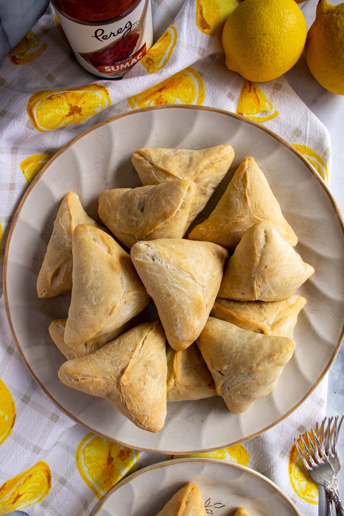 Triangular spinach fatayer pies on a round plate with lemons and sumac spice next to it.
