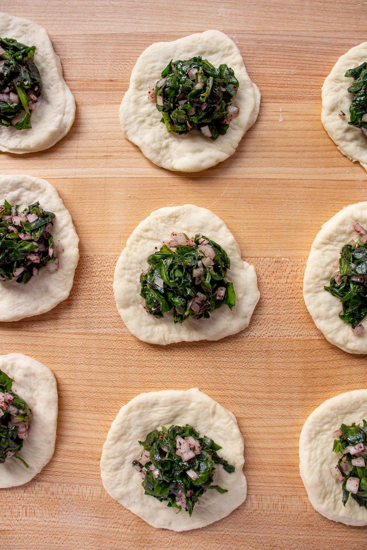 Scoops of spinach filling mounded onto small circles of dough on a wooden board..