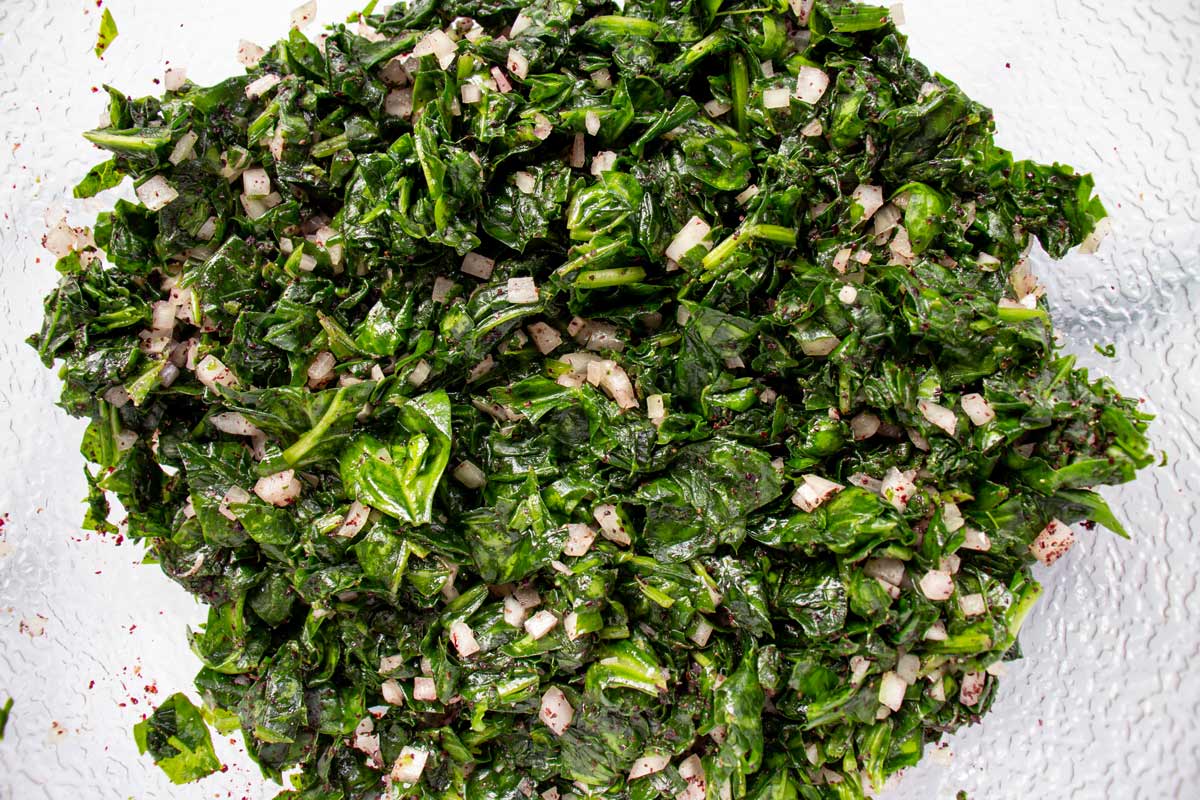 Chopped wilted spinach mixed with chopped onion, sumac, and seasonings in a bowl.
