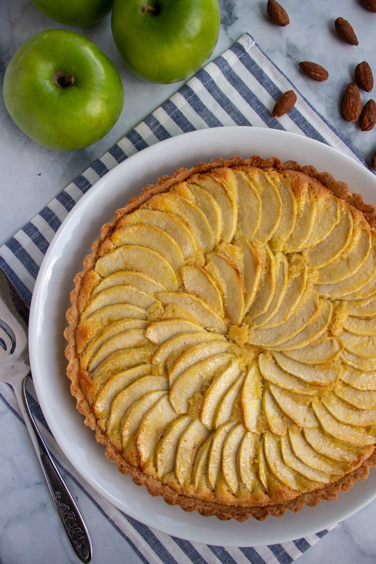 A tart with a spiral of sliced apples on top, with apples and almonds beside it.