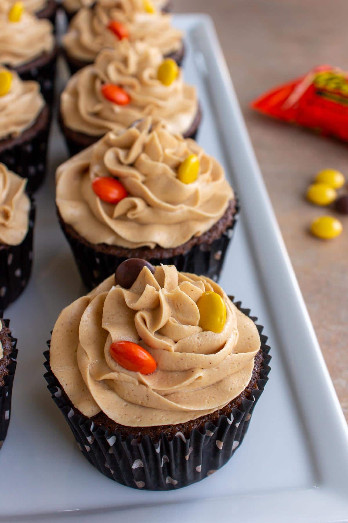A row of Reese's cupcakes topped with Reese's Pieces candies on a white rectangular plate.