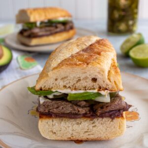 A Mexican pepito sandwich with steak and avocado filling in a baguette on a rustic plate.