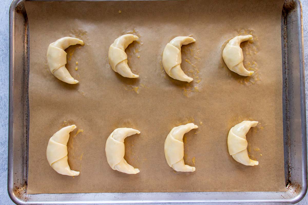 Eight small unbaked croissants brushed with egg wash on a parchment paper lined baking sheet.