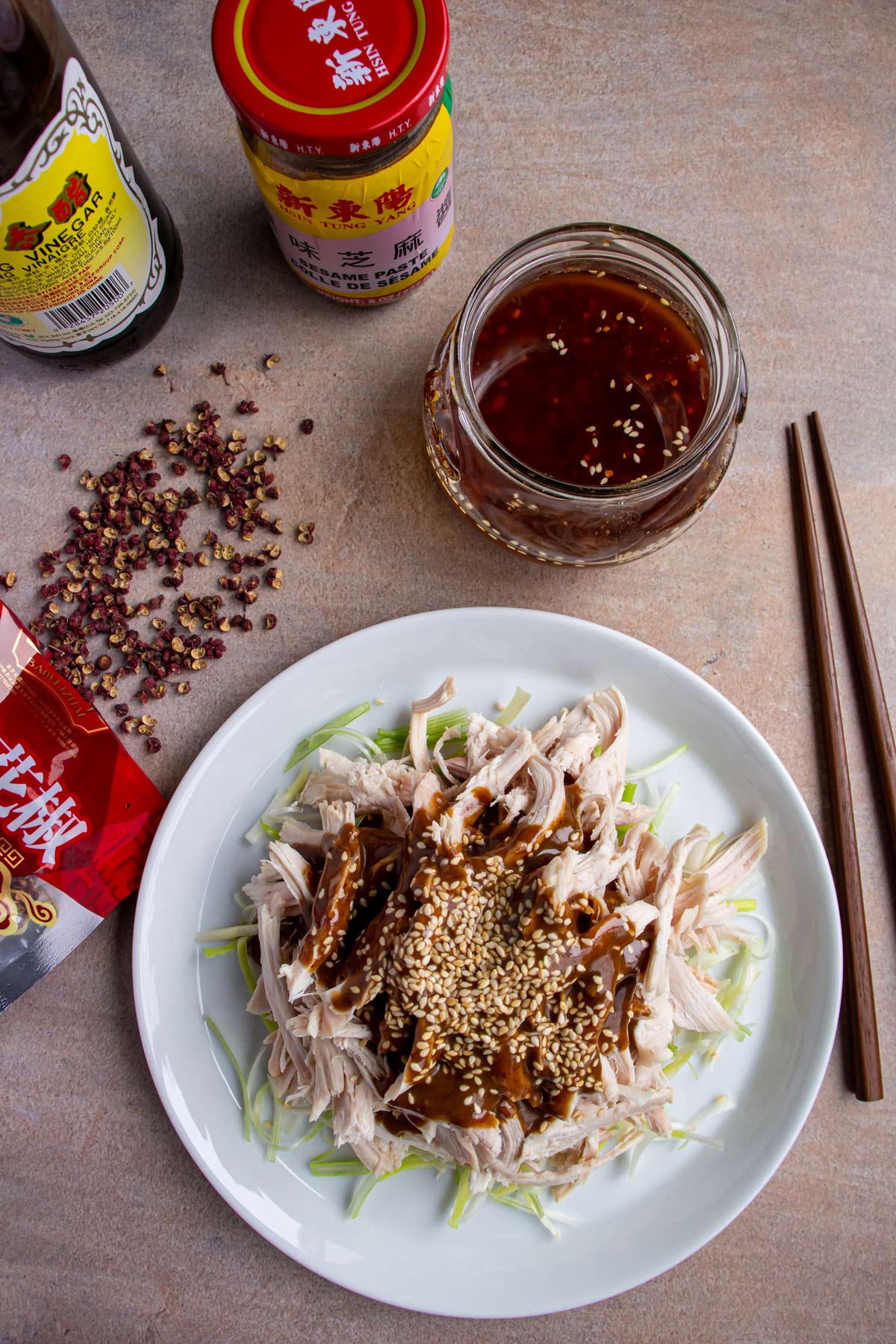 A plate of bang bang chicken surrounded by jars of sauce, Sichuan pepper, and chopsticks.