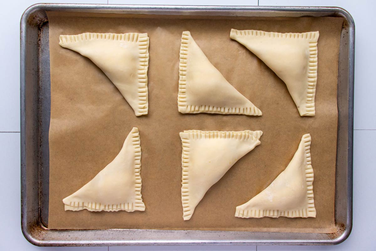 Six unbaked puff pastry triangles with crimped edges on a parchment paper lined baking sheet.