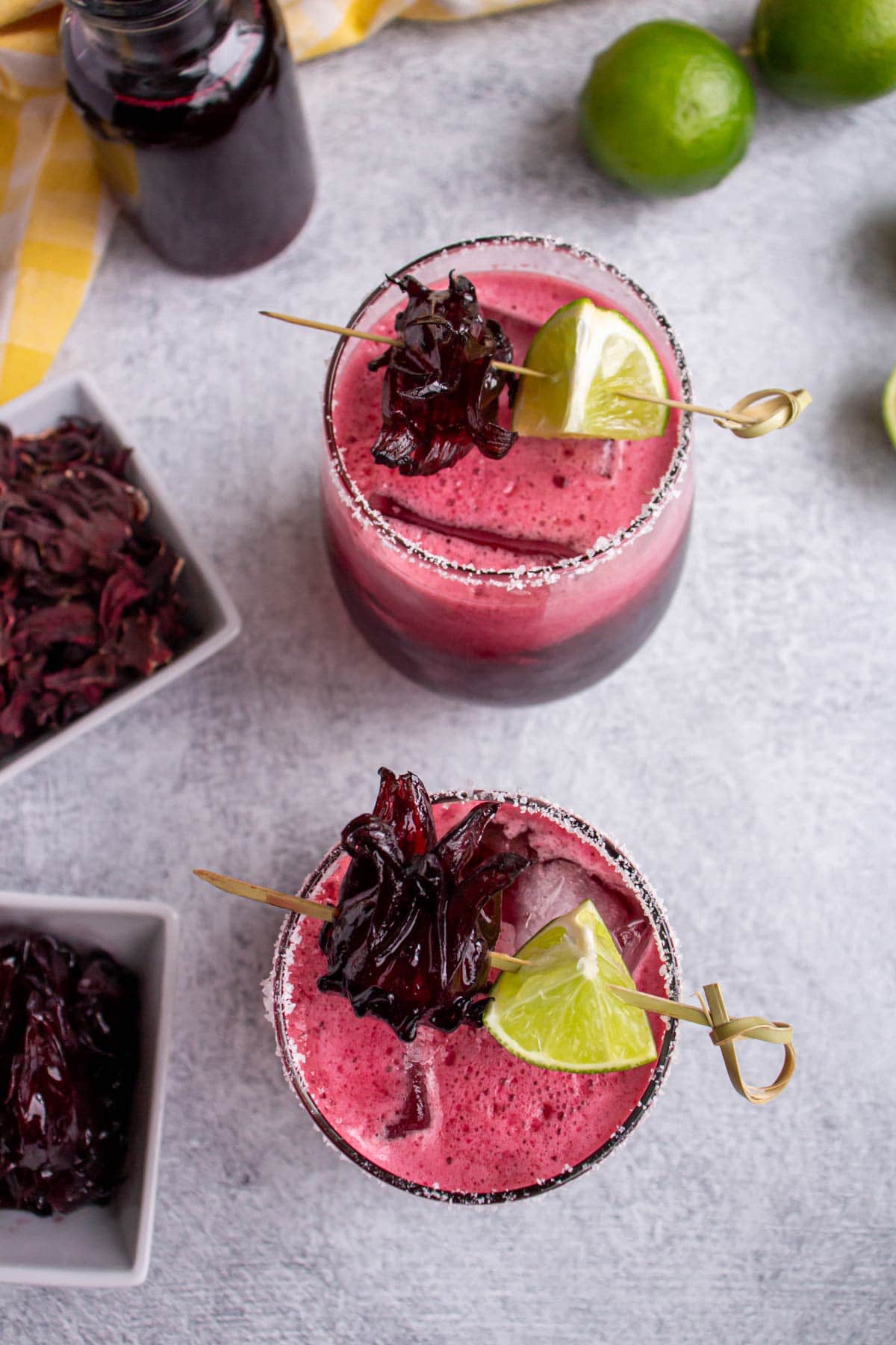 Two pink cocktails with skewered lime wedges and hibiscus flowers on a light grey background.