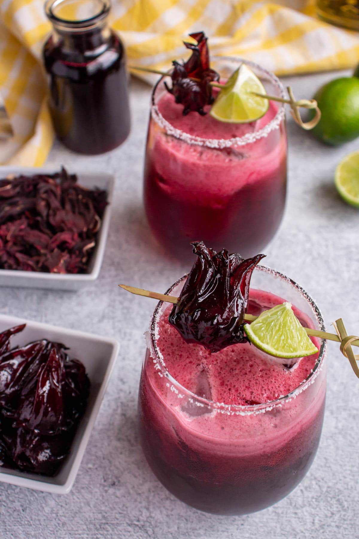 Two hibiscus margaritas garnished with lime and hibiscus flowers with a bottle of syrup and limes.