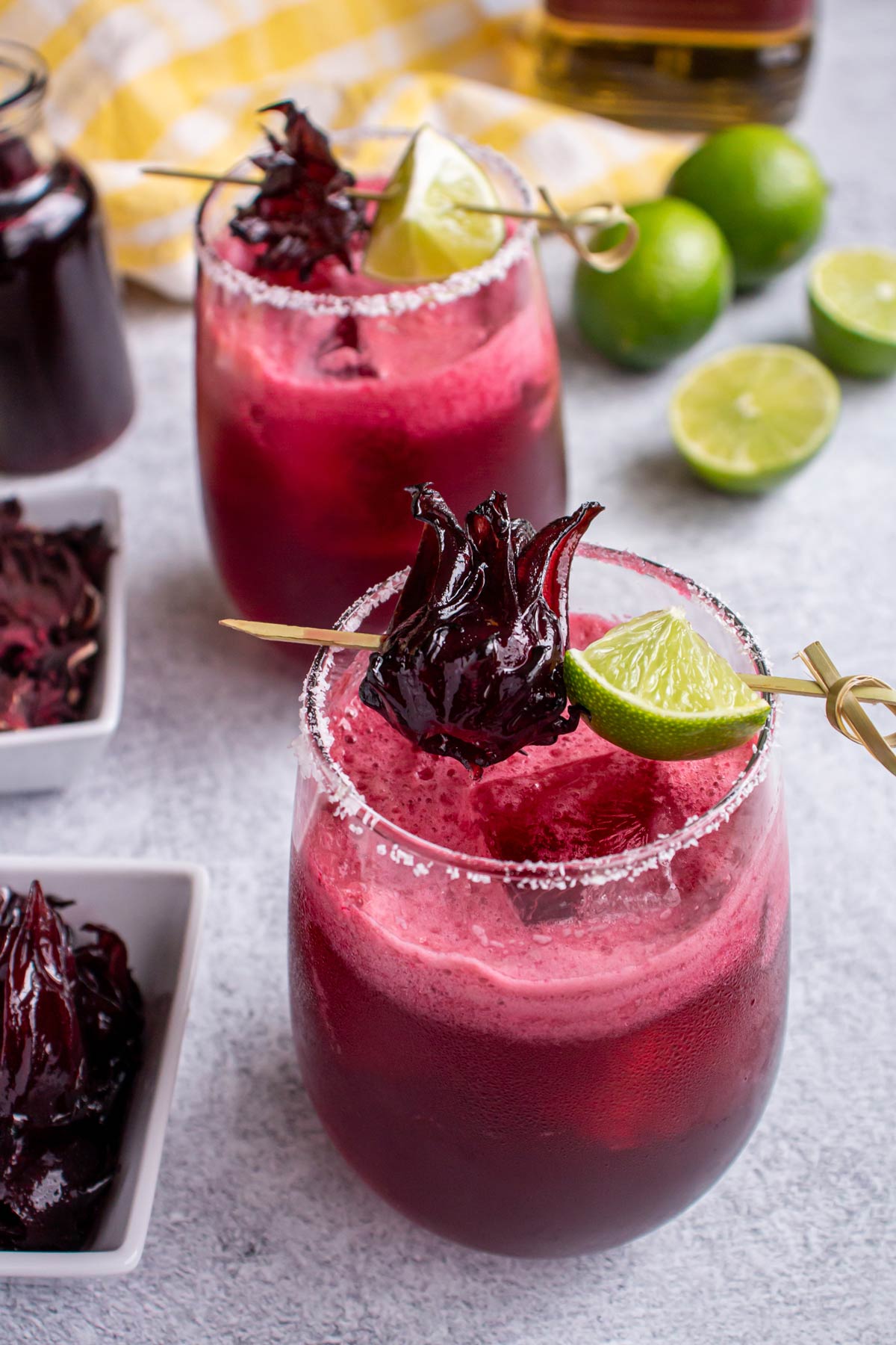 Two bright pink hibiscus margaritas in salt-rimmed glasses garnished with skewered lime and hibiscus flowers.