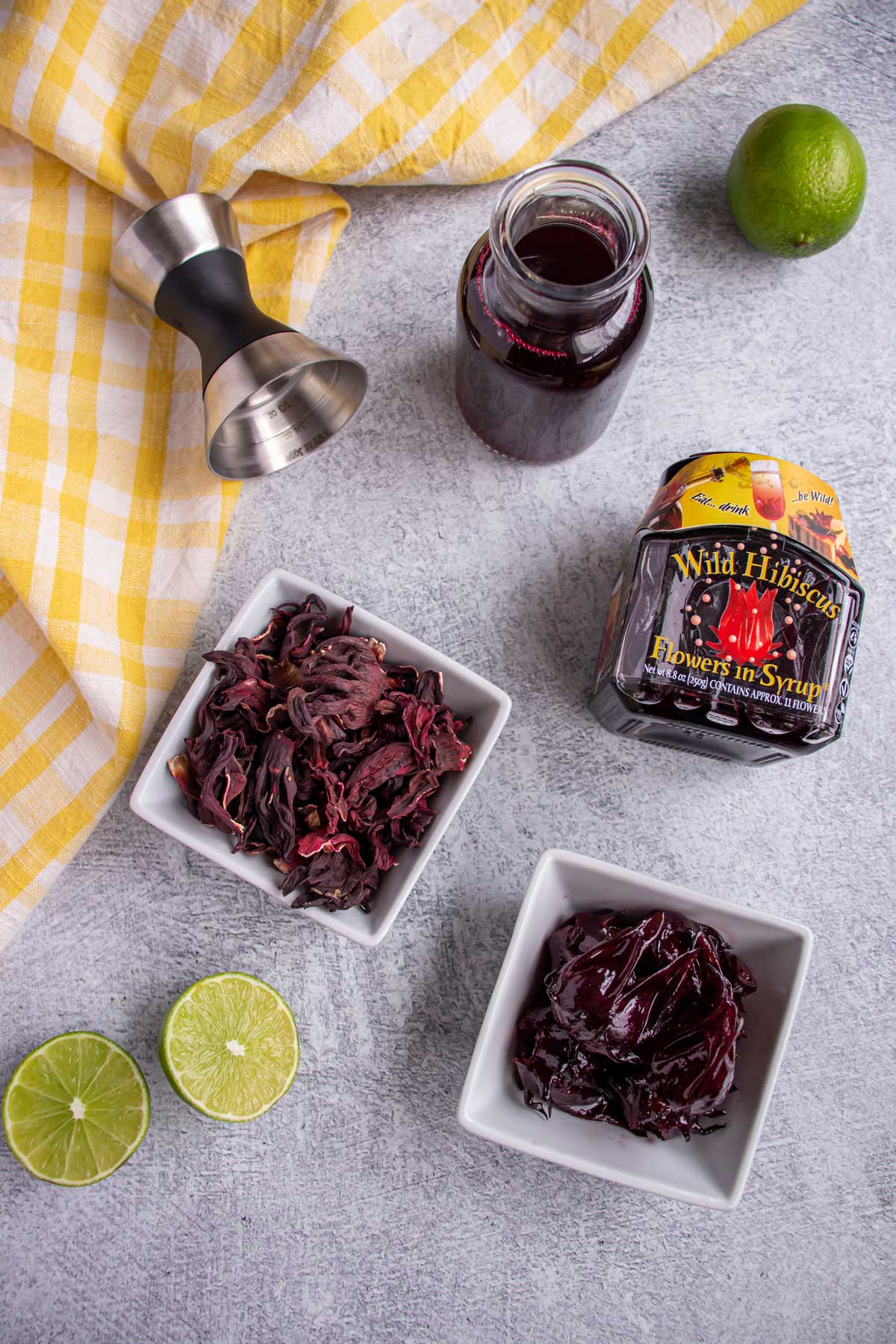 A jigger, small bottle of hibiscus syrup, dried hibiscus flowers and flowers in syrup, and limes.