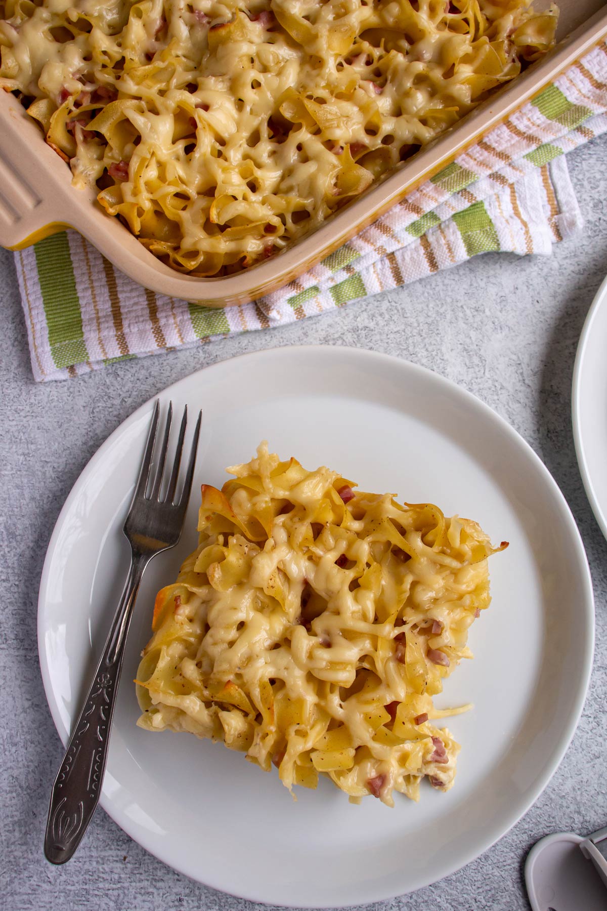 A piece of noodle casserole with a fork on a plate next to a casserole dish.