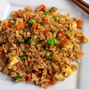 Fried rice with carrots, peas, and scrambled egg on a square plate with wooden chopsticks.
