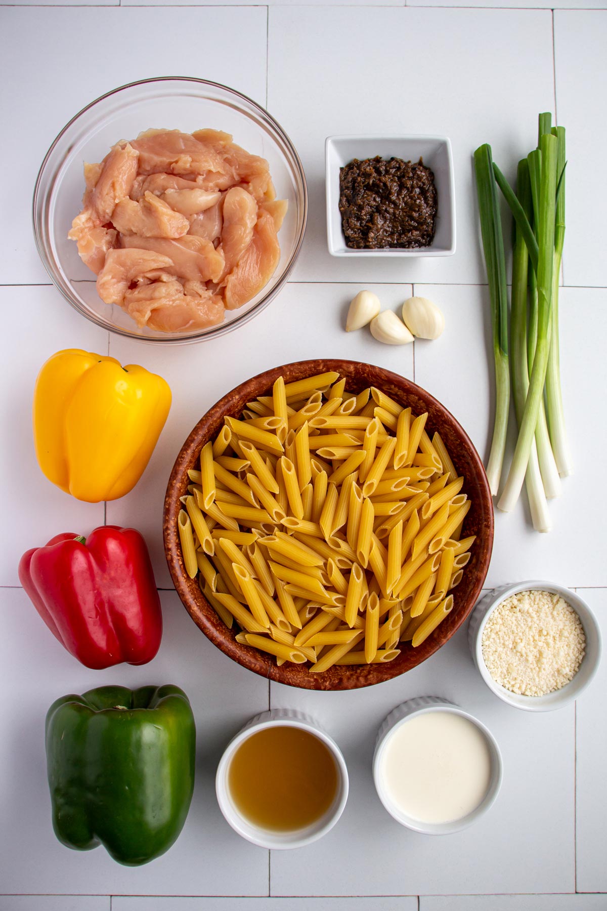 Ingredients for Rasta pasta with jerk chicken on a white tile background.