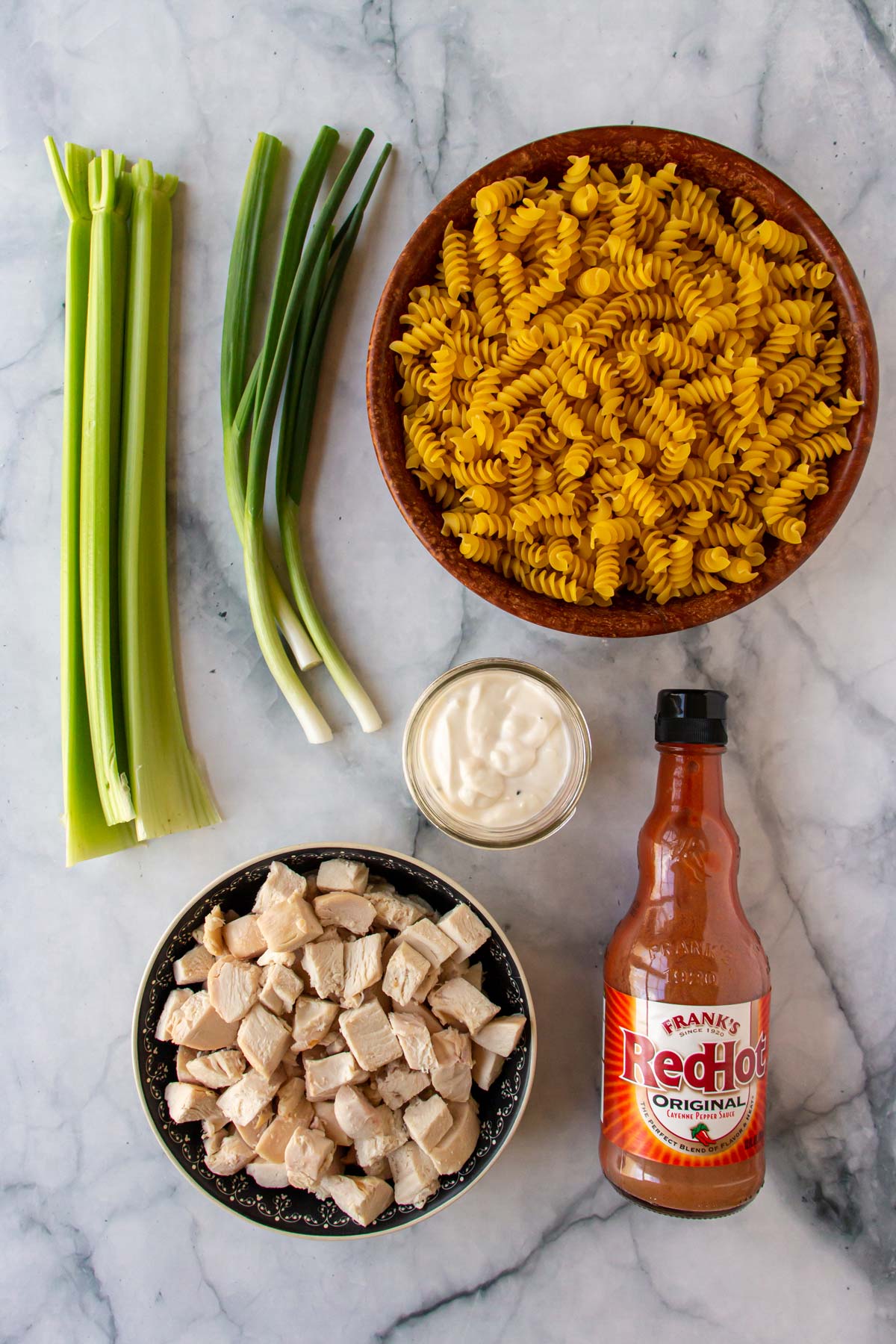 Ingredients for Buffalo chicken pasta salad on a white marble background.