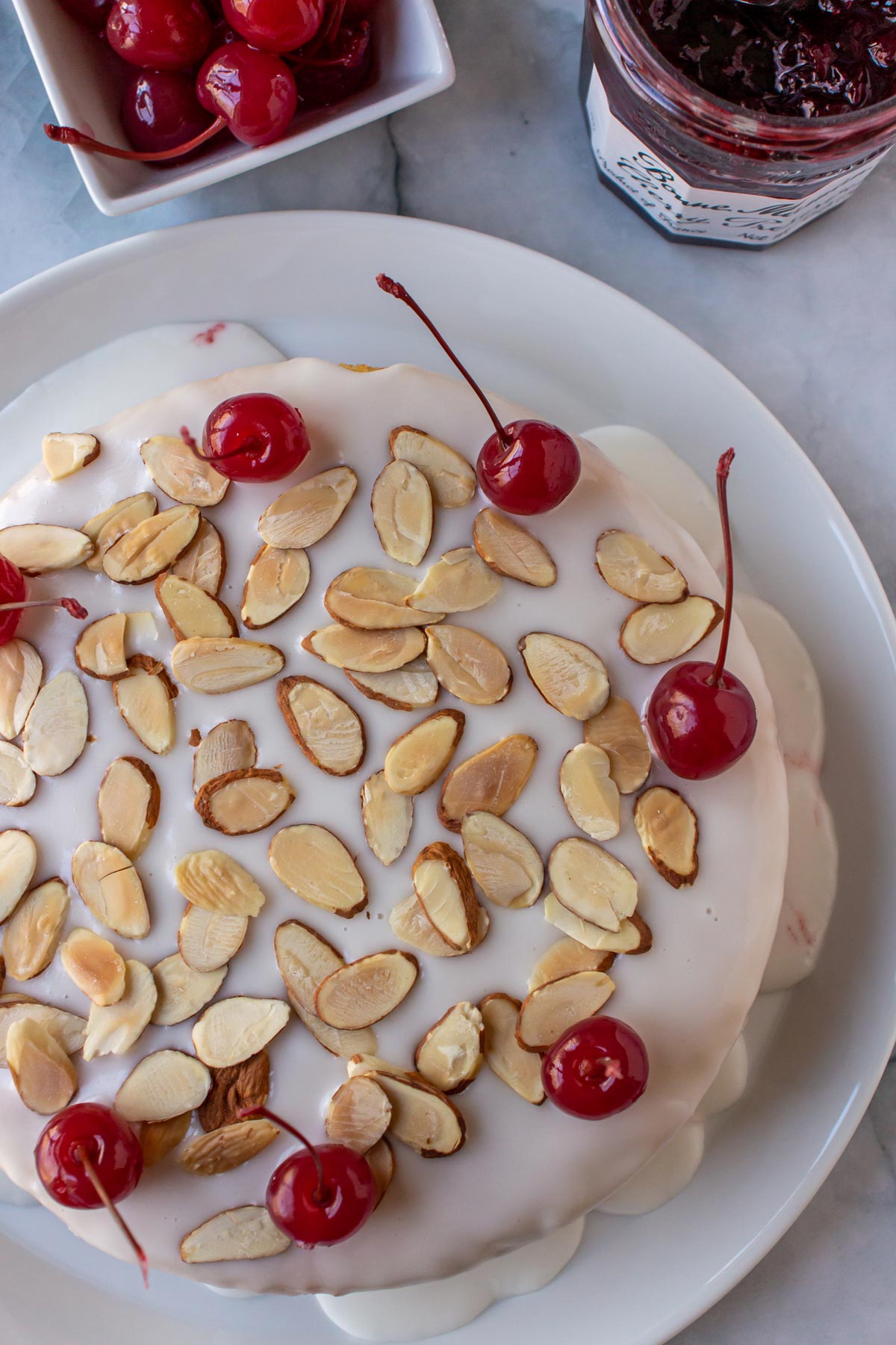 A cake on a white plate topped with white glaze, sliced almonds and maraschino cherries.