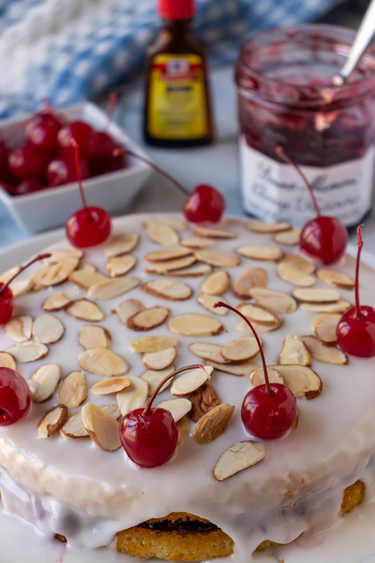 A cherry Bakewell cake garnished with almonds and cherries with a jam jar behind it.