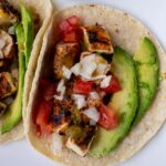 A grilled chicken taco with sliced avocado, chopped tomato and onion on a white plate.
