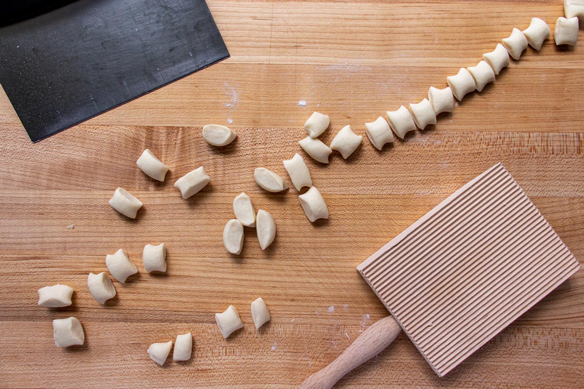 Small pieces of dough, a metal bench scraper and a gnocchi board on a wooden surface.