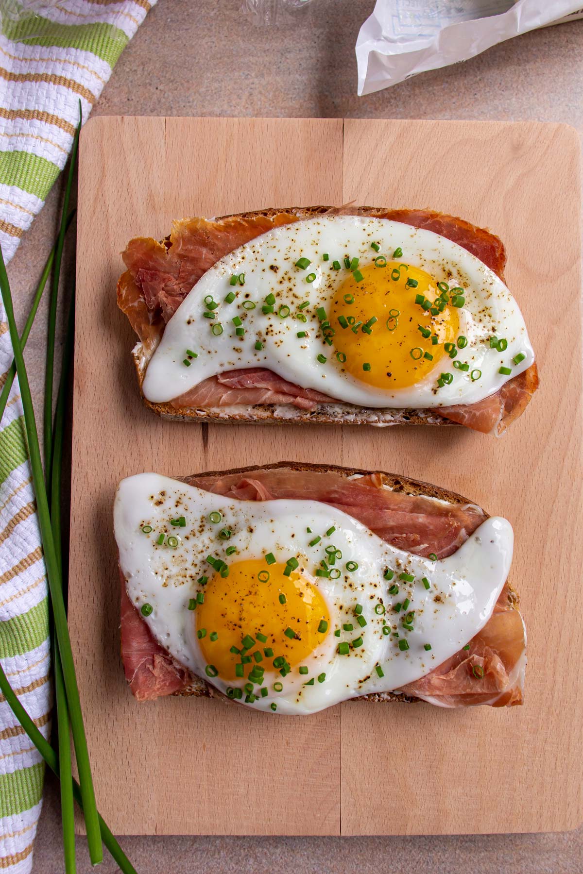Two open-faced cured ham sandwiches with fried egg and chives on a wooden board.