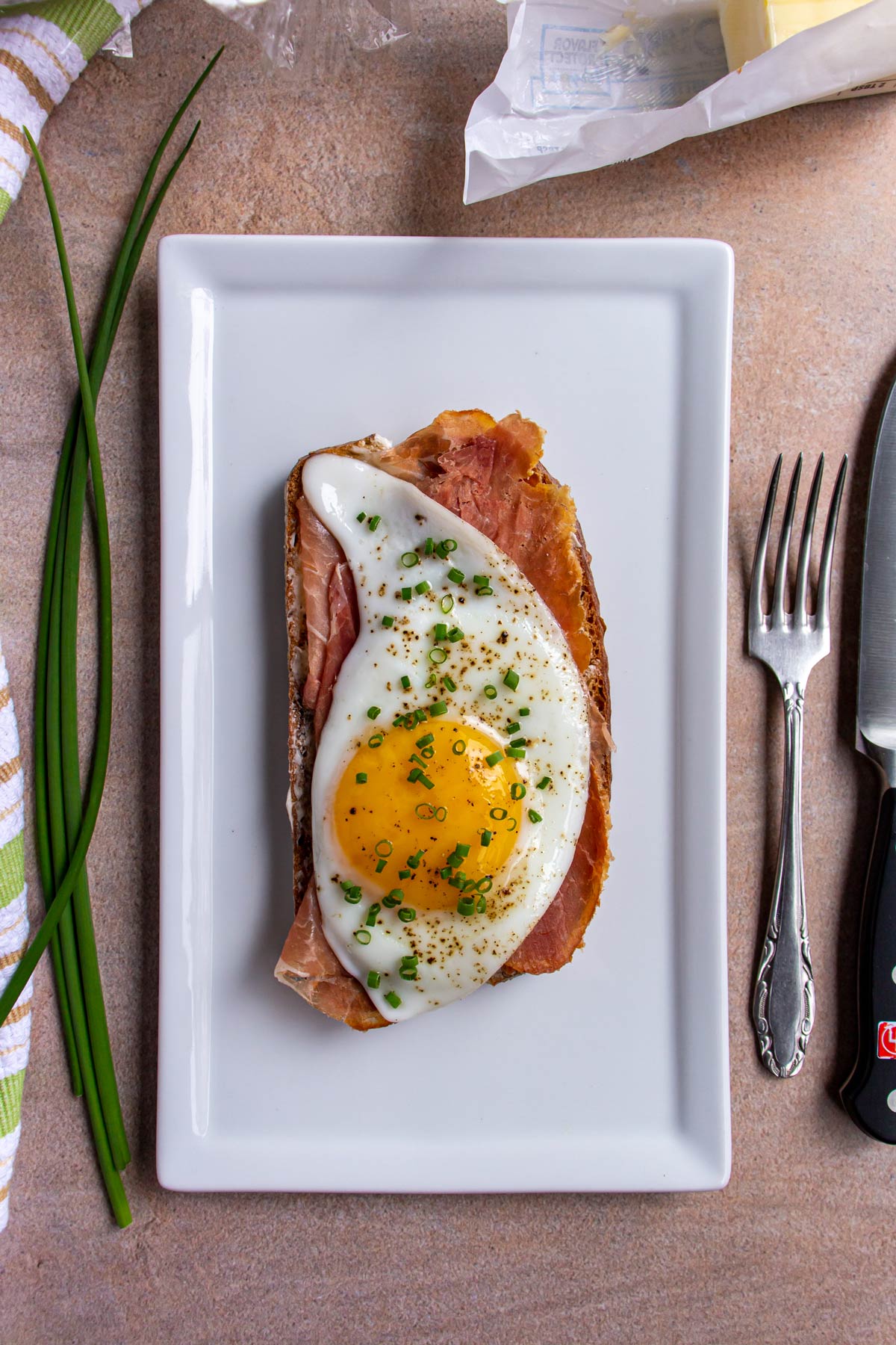 An open-faced sandwich with prosciutto, fried egg, and chopped chives on a white rectangular plate.