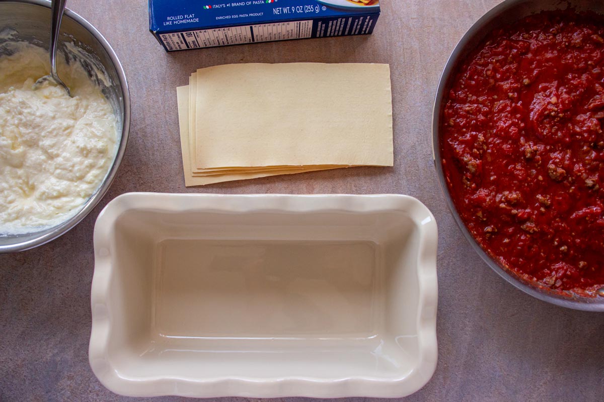 A ceramic loaf pan, a bowl of ricotta filling, dry lasagna sheets, and tomato sauce.