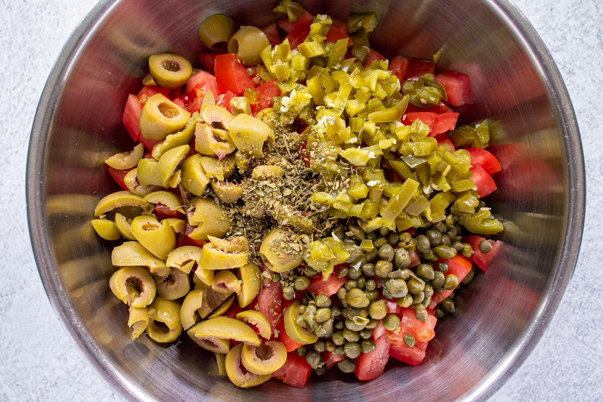 Chopped tomatoes, green olives, pickled jalapenos, capers, and herbs in a metal bowl.