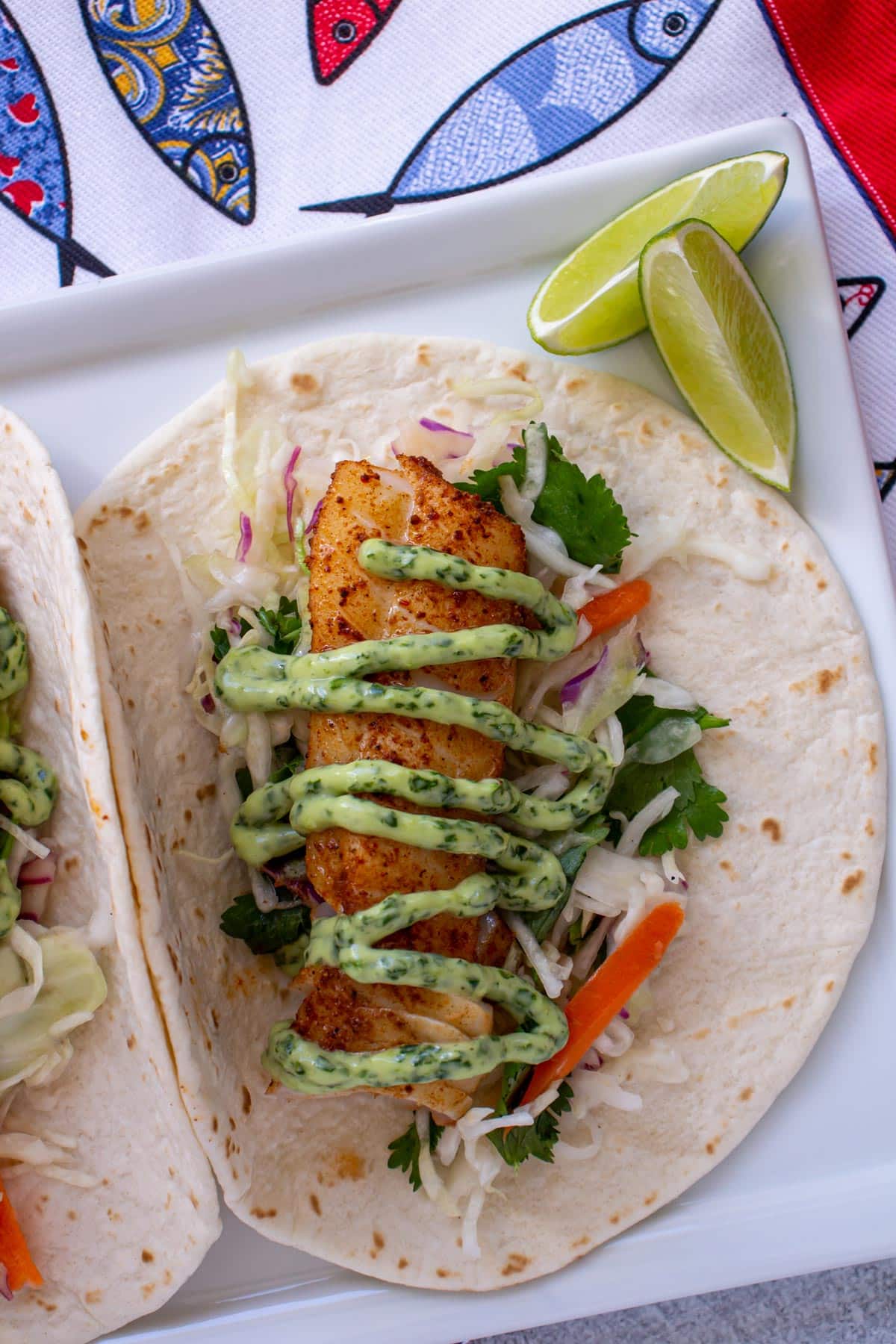 A grilled fish taco with a zig-zag of green cilantro lime mayo and lime wedges.