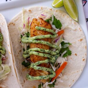 A grilled fish taco with a zig-zag of green cilantro lime mayo and lime wedges.
