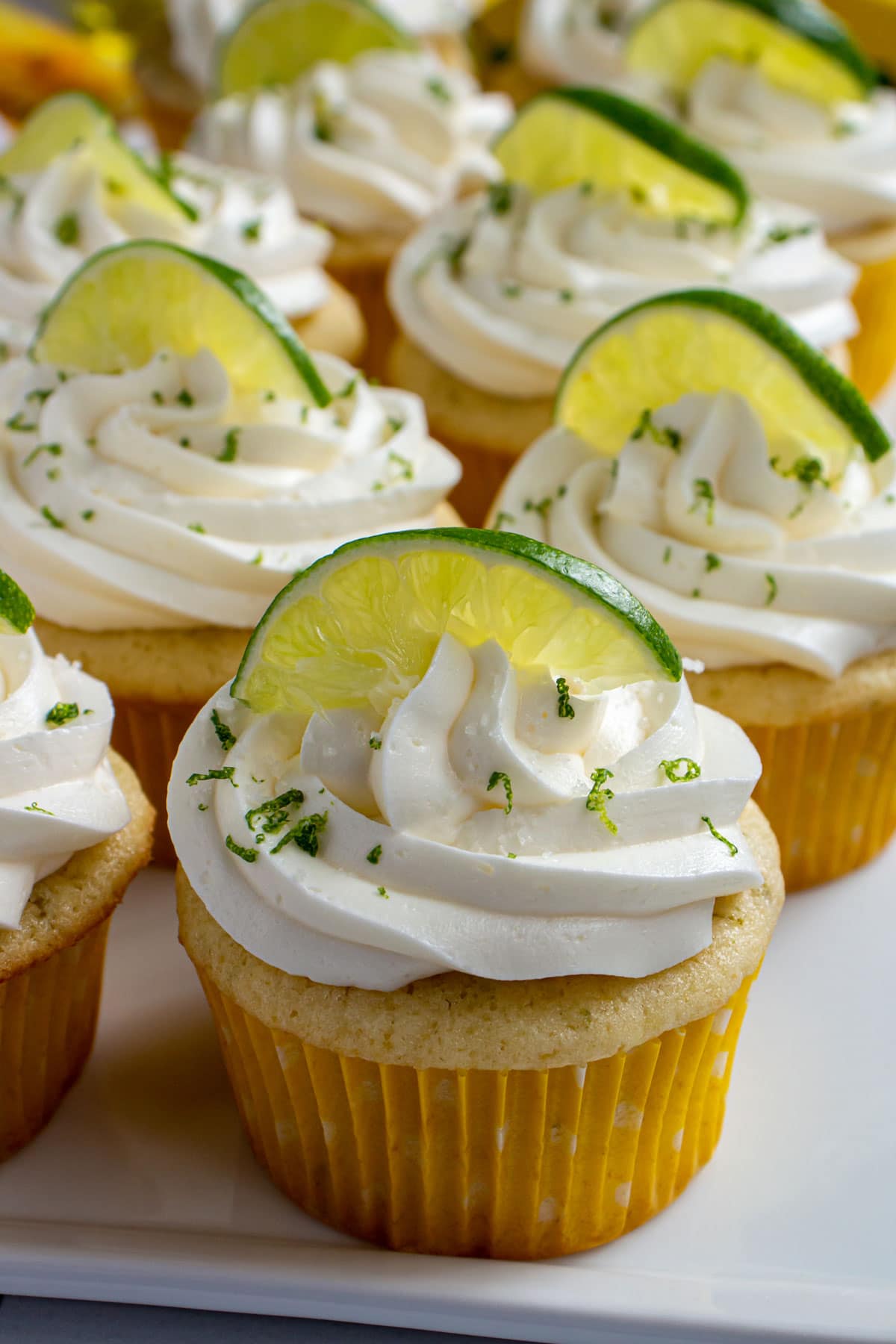 White cupcakes with yellow wrappers, white frosting, and lime slice and zest garnish.