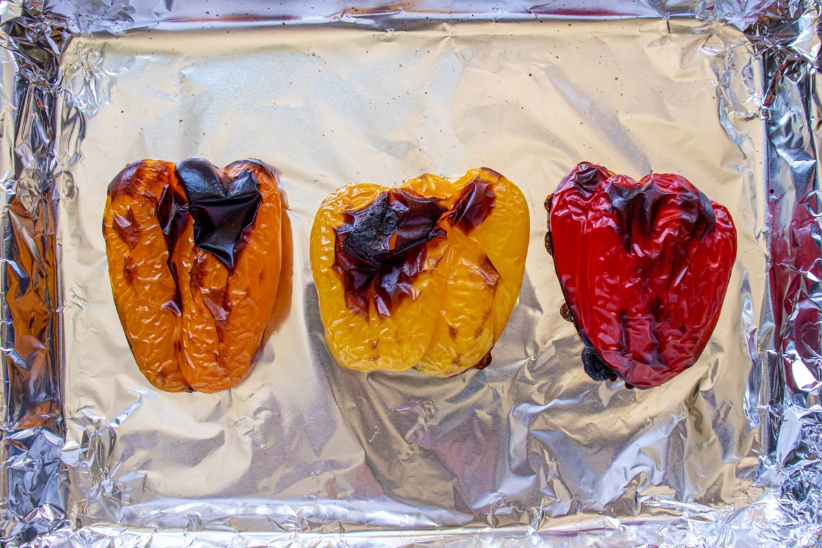 Orange, yellow, and red bell pepper halves roasted with blistered skin on a foil lined pan.