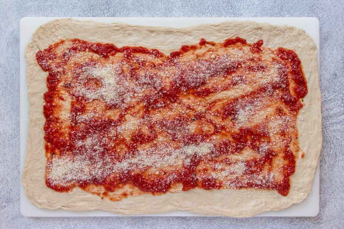 A rectangle of stretched out pizza dough topped with tomato sauce and grated cheese.