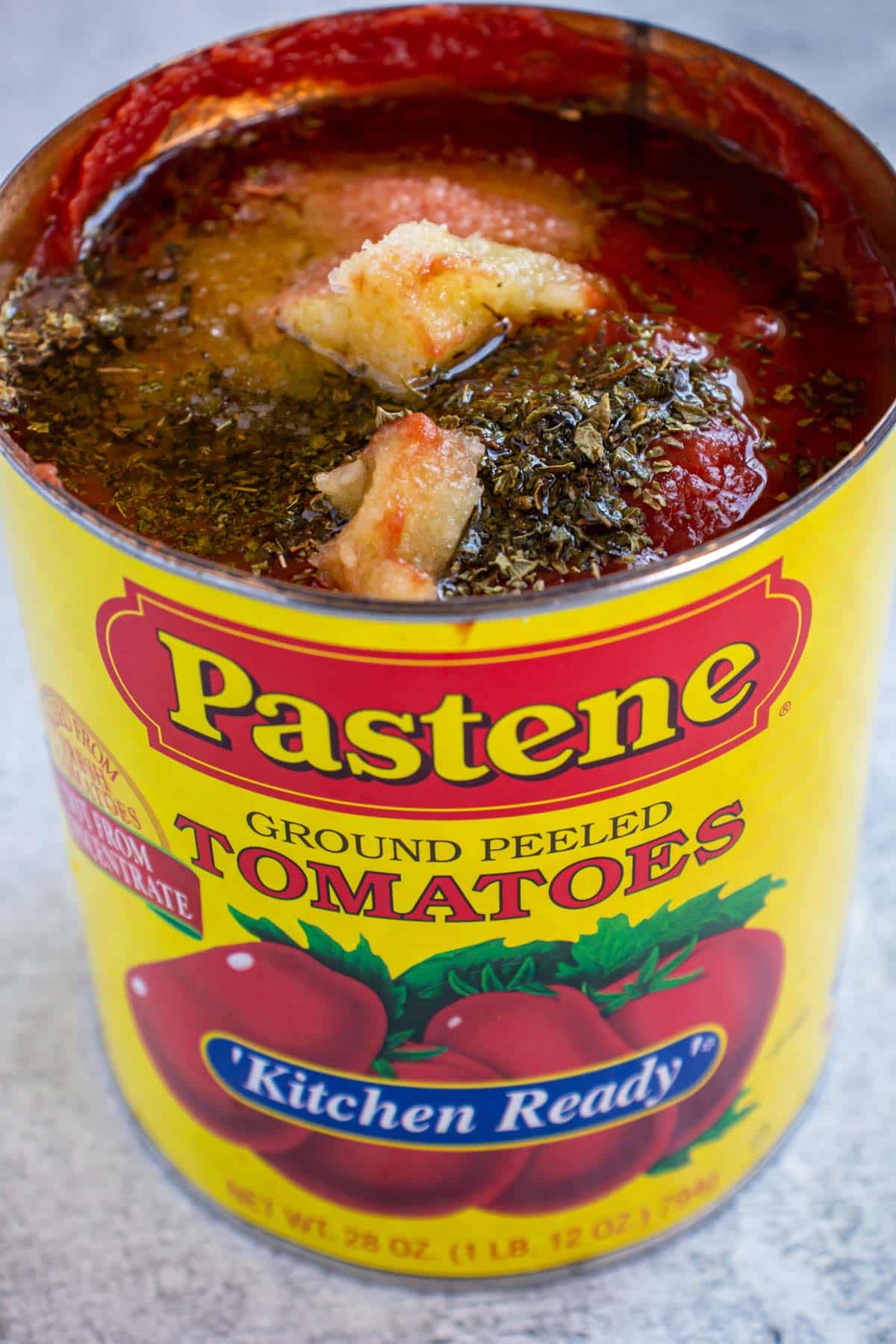 An open can of Pastene ground peeled tomatoes with dried oregano and crushed garlic on top.