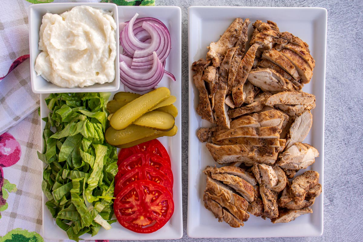 A platter of sliced chicken and a platter of toppings including veggies, pickles, and garlic sauce.