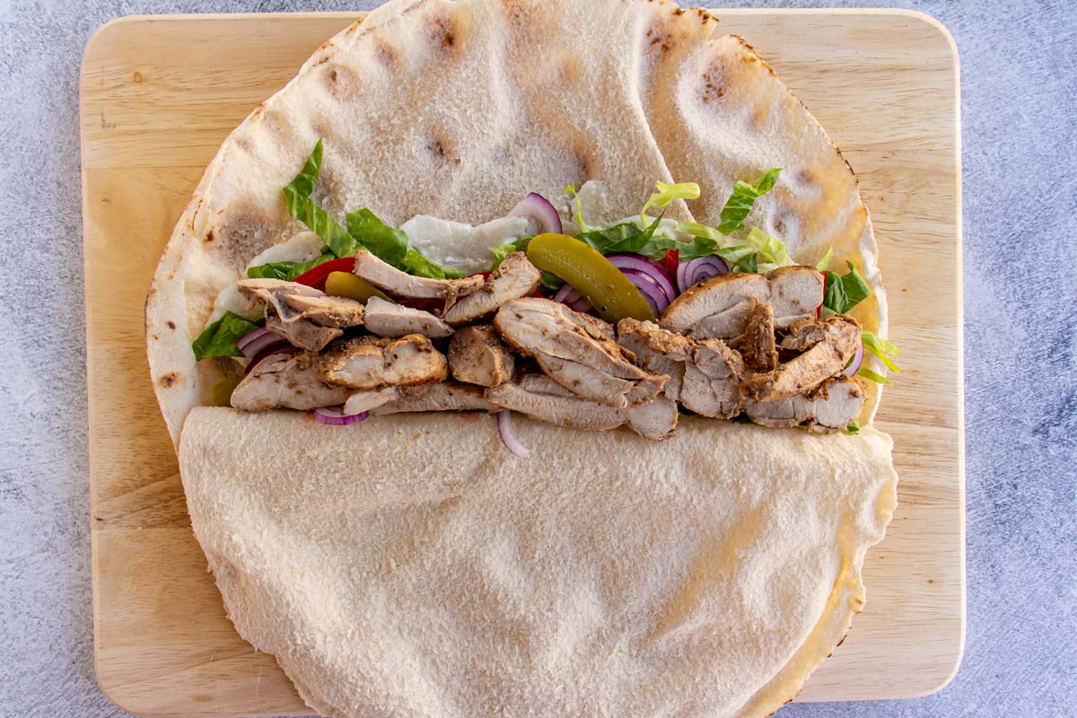 A half-opened pita flatbread filled with lettuce, tomato, red onions, pickles, and sliced chicken.