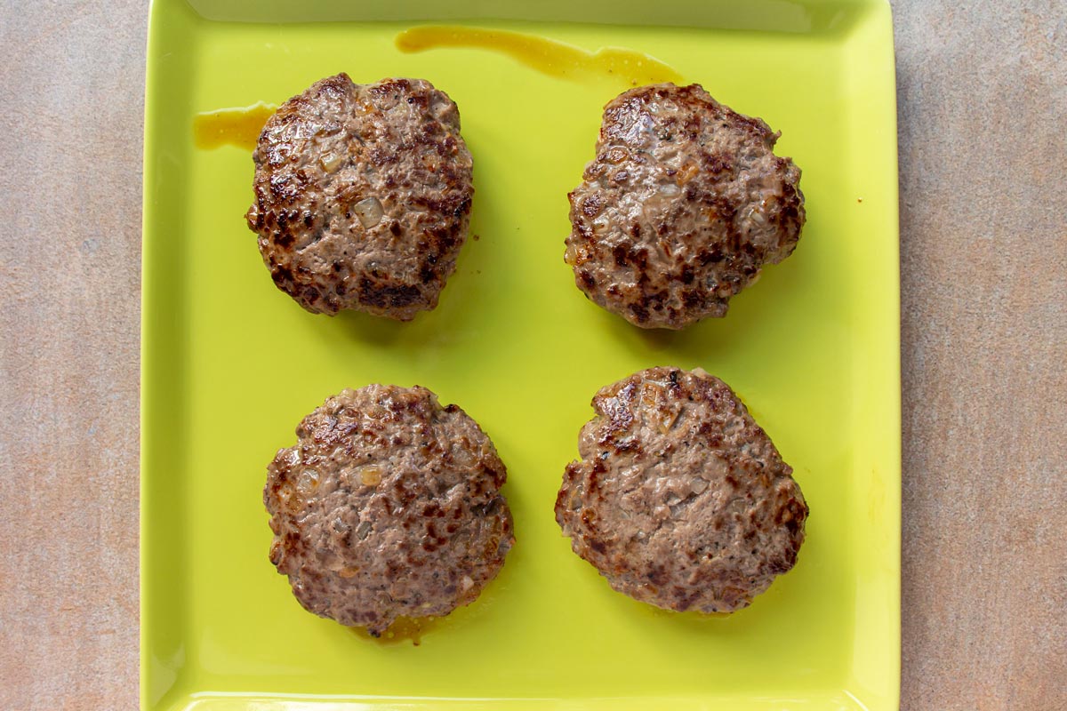 Four cooked beef burger patties on a light green square plate.